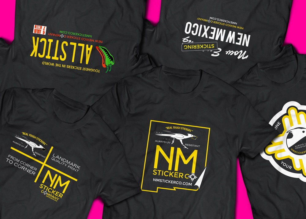 A pile of black shirts on a bright pink background. The designs are varying images of roadrunners and taglines done for New Mexico Sticker Company. Images say "Shine is On Your Side", "Allstick", "Now Stickering New Mexico", and "Landmark Print from Corner to Corner".