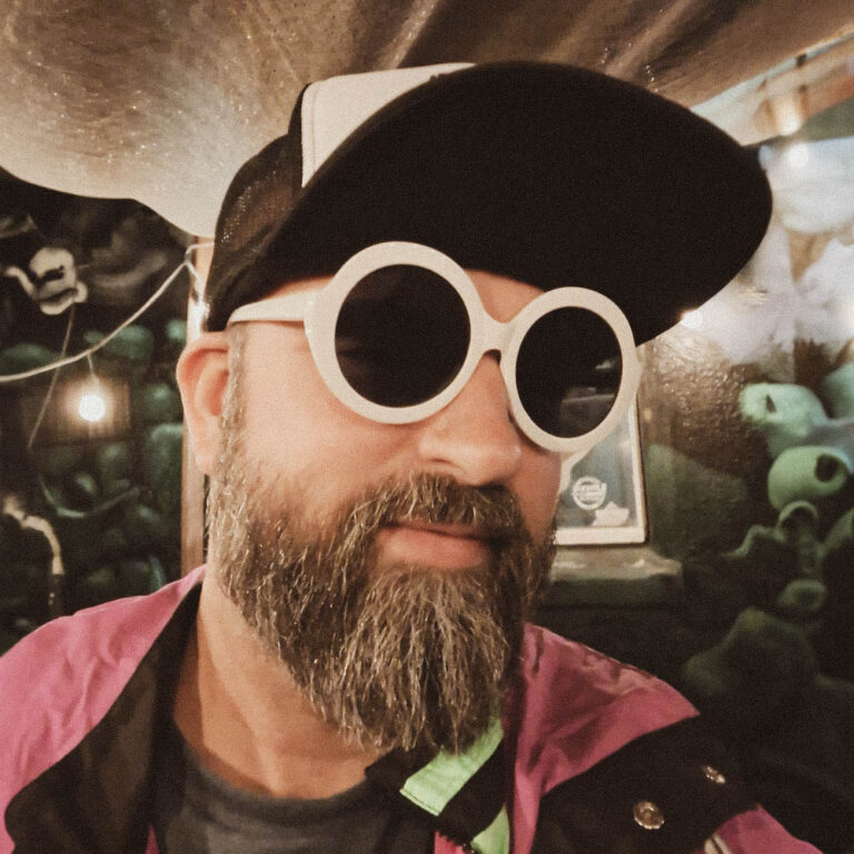 Man in a beard dressed as Rick Nielson of Cheap Trick. He is wearing a flip bill baseball cap, white circle sunglasses, and a neon green and yellow jacket in front of a black and green abstract mural.
